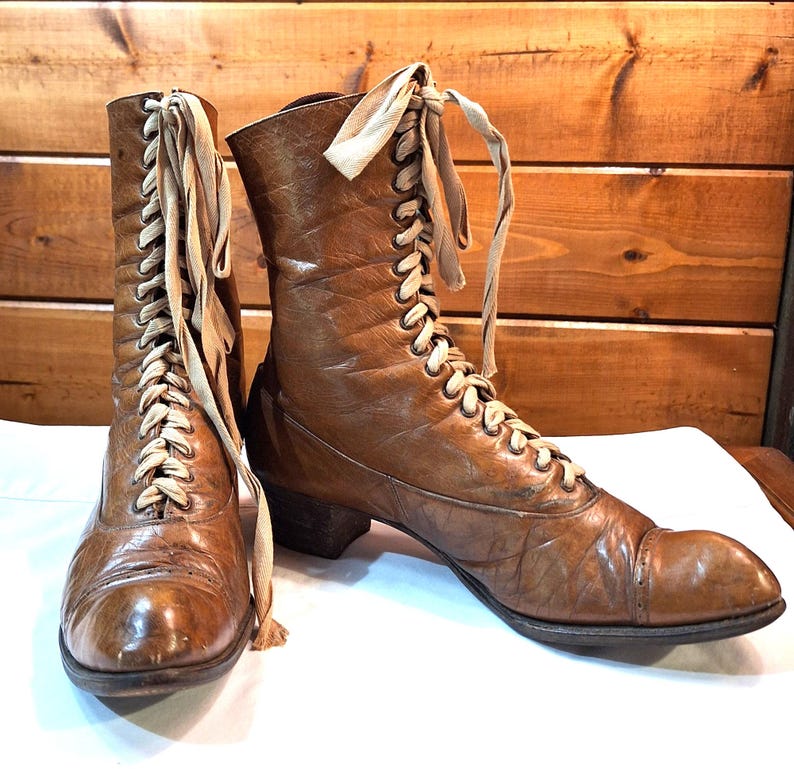 A pair of brown boots with tan laces that are Victorian style with long narrow length and above the ankle height.