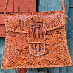Close up image of a square snakeskin handbag with a tab closure in the shape of a snake's head in front. The colors are chestnut and black and the purse is structured.