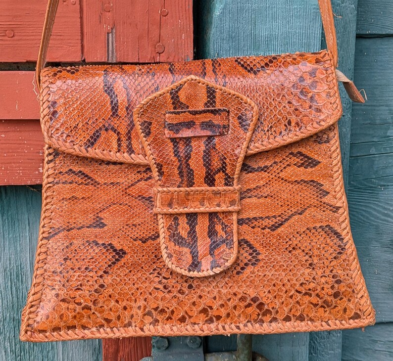 Close up image of a square snakeskin handbag with a tab closure in the shape of a snake's head in front. The colors are chestnut and black and the purse is structured.