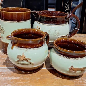 Può includere: Un set di quattro tazze da tè in ceramica marrone e bianca con scene di caccia. Le tazze hanno una smaltatura marrone con una fascia bianca e una scena di caccia al centro. Le tazze sono tutte di dimensioni e forme diverse.