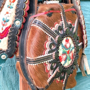 Puede incluir: Un bolso de cuero y piel marrón con intrincados bordados florales y detalles decorativos de cuero negro. El bolso presenta un diseño detallado con patrones florales rojos, blancos y verdes, y es probablemente una pieza de arte tradicional o popular.