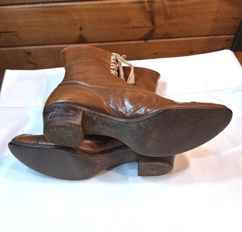 Bottom view of the soles of a pair of ladies brown leather Victorian boots with tan laced fronts. The soles have dark leather with tan stitching .