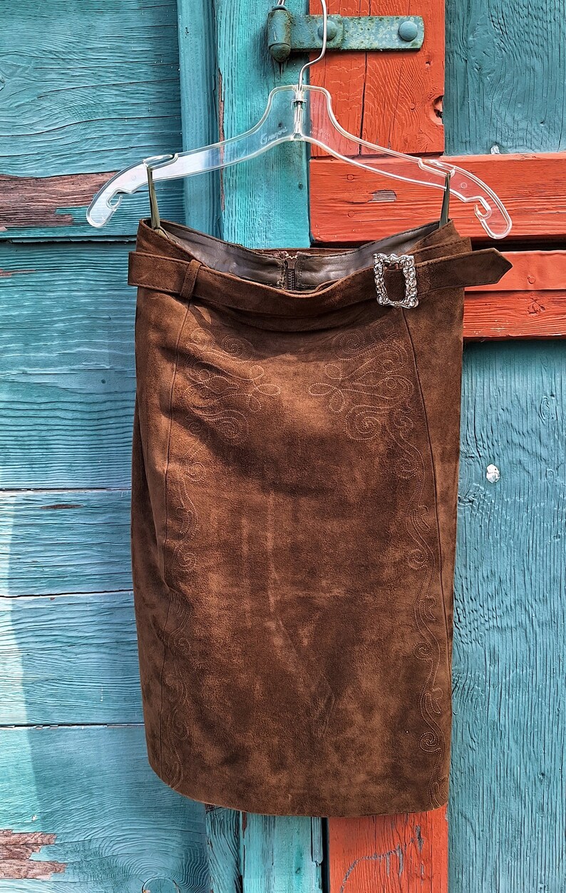 A brown suede skirt with a decorative embroidered design. The skirt has a belt with a silver buckle and a zipper closure.