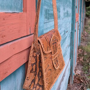 Right side view of a structured snakeskin hand back that is chestnut and black. the bottom is slightly flared and it has a front tab closure and a long shoulder strap.