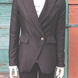 Pode incluir: Um blazer preto com um único botão dourado, combinado com calças de veludo cotelê pretas. O blazer apresenta lapelas entalhadas e um corte sob medida. Uma camisa branca com babados é visível por baixo.