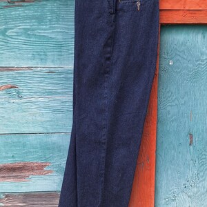 Dark blue denim riding jods with a cuffed hem hanging on a wooden hanger. Image is showing the jods from the side showng the side pocket and width of the leg