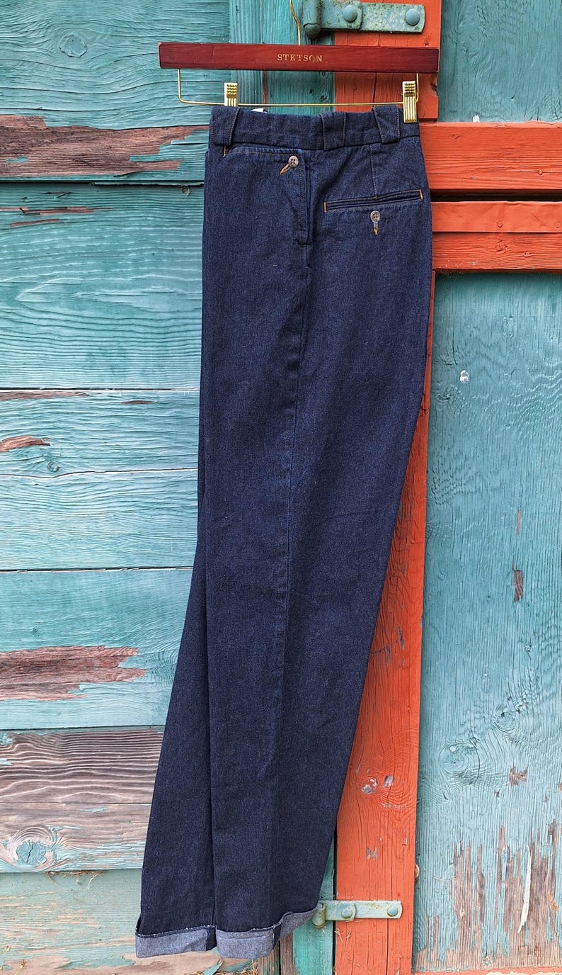Dark blue denim riding jods with a cuffed hem hanging on a wooden hanger. Image is showing the jods from the side showng the side pocket and width of the leg
