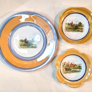 Puede incluir: Tres platos decorativos con una escena de caza. El plato más grande tiene un borde azul y naranja. Los platos más pequeños tienen un borde dorado. Cada plato presenta una imagen central de una persona a caballo con perros.