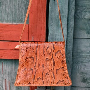 Backside image of a structured snakeskin handbag. The purse is square with a flared bottom and black and chestnut colors with a long shoulder strap.