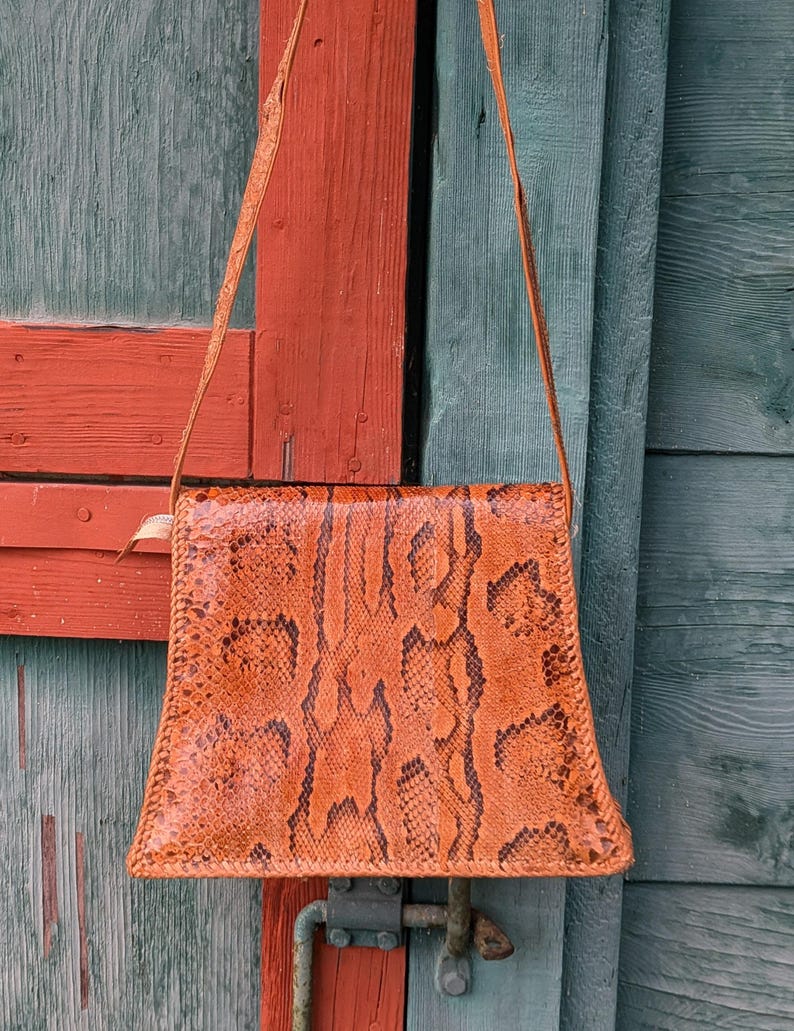 Backside image of a structured snakeskin handbag. The purse is square with a flared bottom and black and chestnut colors with a long shoulder strap.