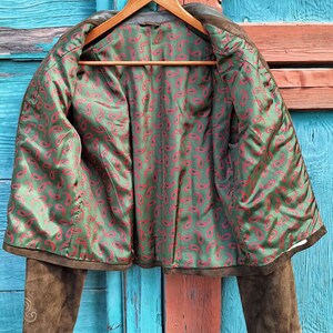 A brown suede jacket with a green and red paisley lining. The jacket is unbuttoned and the lining is visible. The jacket is hanging on a wooden hanger against a blue and orange wooden background.