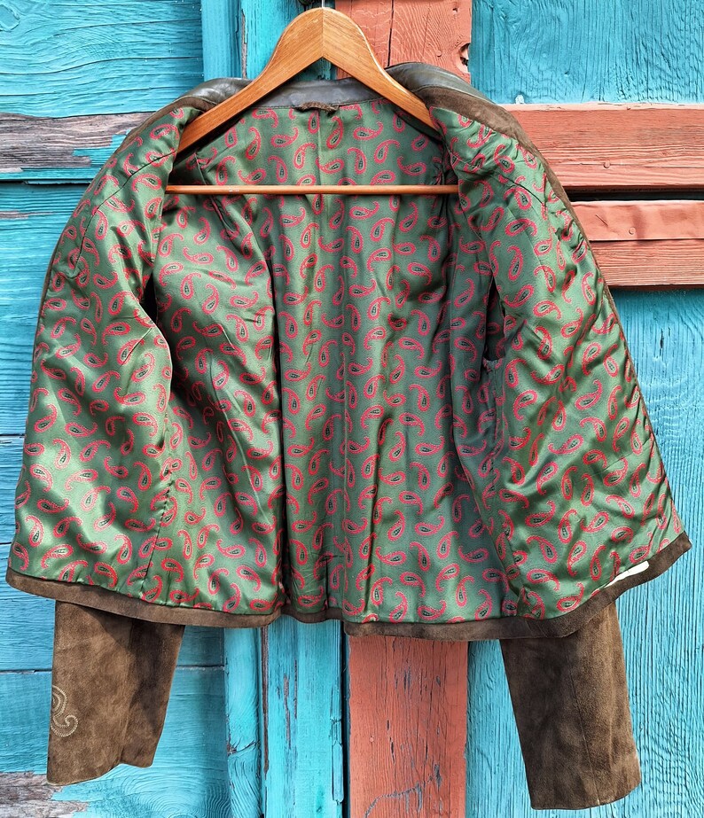 A brown suede jacket with a green and red paisley lining. The jacket is unbuttoned and the lining is visible. The jacket is hanging on a wooden hanger against a blue and orange wooden background.