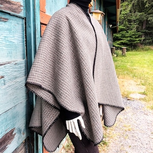 May include: A beige and black houndstooth patterned poncho featuring a black stitched edge trim. The garment is draped over a black turtleneck sweater and dark trousers, showcasing a loose, open-front silhouette with a textured fabric finish.