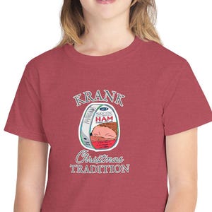 May include: A red t-shirt with a graphic of a ham can and the text "Krank Christmas Tradition" in white and teal. The shirt is made of a soft material, suitable for casual wear. The design is festive.