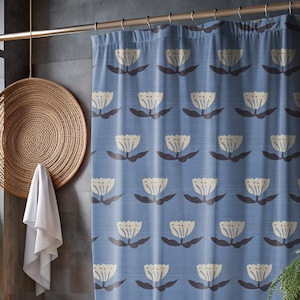 Peut inclure: Rideau de douche bleu avec un motif floral répétitif crème et marron. La salle de bain comprend une baignoire blanche, un panier en osier, un petit tabouret en bois et une plante en pot. Le rideau est suspendu à une tringle dorée.