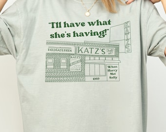 以下が含まれることがあります： 緑色のグラフィックが特徴のライトグリーンのTシャツ。Katz's Delicatessenのイラストと「I'll have what she's having!」と「When Harry Met Sally」の文字がプリントされています。カジュアルなスタイルです。