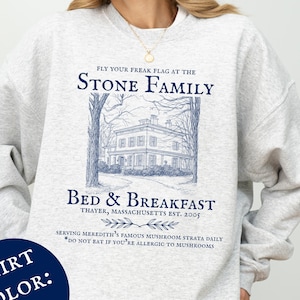 May include: Ash-colored sweatshirt with a blue illustration of a house and text that reads "STONE FAMILY BED & BREAKFAST". The text also includes "FLY YOUR FREAK FLAG AT THE" and "SERVING MEREDITH'S FAMOUS MUSHROOM STRATA DAILY".