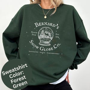 May include: A forest green crewneck sweatshirt featuring a white graphic of a snow globe with a cabin inside. The text reads: Bernard's Snow Globe Co., North Pole, Est. 1994, Handcrafted, Magical, Shake Responsibly, surrounded by small white dots.