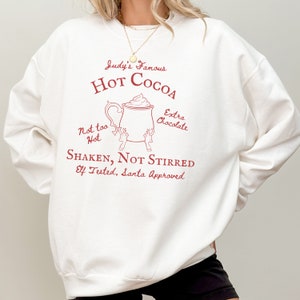 May include: White sweatshirt with a red graphic design of a mug of hot cocoa with the text "Judy's Famous Hot Cocoa", "Not too Hot", "Extra Chocolate", "Shaken, Not Stirred", and "Elf Tested, Santa Approved".