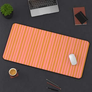 Op de afbeelding: Een roze en oranje gestreepte bureau mat met een witte computermuis erop. De mat ligt op een zwart oppervlak met een kopje koffie, potloden en een laptop.