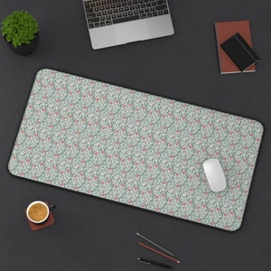 May include: A white desk mat with a floral pattern in shades of green and pink. The mat is on a black surface with a laptop, a cup of coffee, a mouse, and pencils.