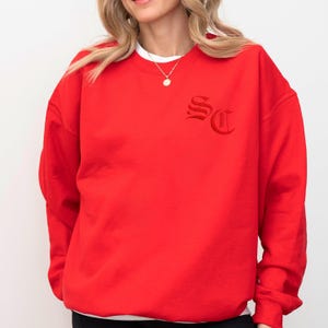 May include: A bright red sweatshirt with the embroidered letters "SC" in a gothic font on the upper left chest. The sweatshirt has a crew neck and long sleeves. A white undershirt is visible at the neckline.