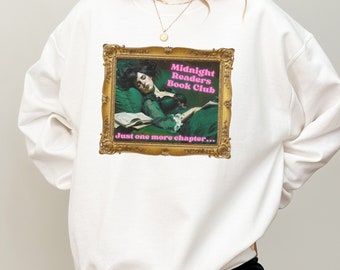 Puede incluir: Sudadera blanca con una imagen enmarcada en oro de una mujer durmiendo con un libro. El texto "Midnight Readers Book Club" y "Just one more chapter..." está impreso en rosa.