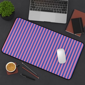Op de afbeelding: Een roze en blauwe gestreepte muismat met een witte computermuis erop. De muismat ligt op een zwart oppervlak met een laptop, een kopje koffie en potloden.