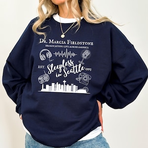 Puede incluir: Sudadera azul marino con un diseño gráfico blanco con el texto "Dr. Marcia Fieldstone Broadcasting Live Across America" y "Sleepless in Seattle" con un horizonte, auriculares, un micrófono, una radio y un teléfono de disco.