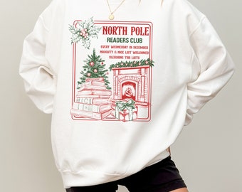 May include: White sweatshirt with a red and green illustration of a fireplace, a stack of books, and a Christmas tree. The text "North Pole Readers Club" is written in red above the illustration. The text "Every Wednesday in December" is written in red below the illustration. The text "Naughty & Nice List Welcomed" is written in red below the text "Every Wednesday in December". The text "Sleighing for Lists" is written in red below the text "Naughty & Nice List Welcomed".