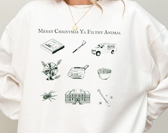 Może przedstawiać: Biała bluza z zielonym nadrukiem graficznym, na którym widnieje napis "Merry Christmas Ya Filthy Animal". Grafika zawiera obrazy domu, pająka, wentylatora, szczoteczki do zębów, furgonetki, miski z płatkami śniadaniowymi, łopaty i kasety.