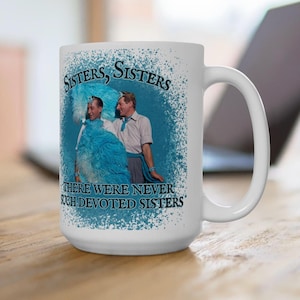 May include: White ceramic mug featuring a vintage image of two people with the text "Sisters, Sisters, There Were Never Such Devoted Sisters." The image is surrounded by a blue and white design. The mug has a large handle.