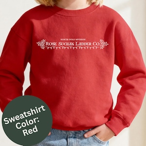 May include: A red crewneck sweatshirt featuring white text that reads North Pole Division Rose Suchak Ladder Co. The design is flanked by holly leaf illustrations and includes a string of decorative holiday lights underneath the main text.