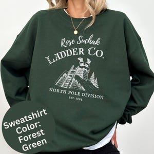 May include: A forest green crewneck sweatshirt featuring a white graphic print of a chimney with legs sticking out, a ladder, and the text Rose Suchak LADDER CO. NORTH POLE DIVISION EST. 1994. The design includes a small pine tree illustration.