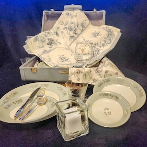 Puede incluir: Una maleta azul vintage con un mantel de flores y un juego de platos con estampado floral blanco y azul, un portavelas de vidrio y una copa de vino.
