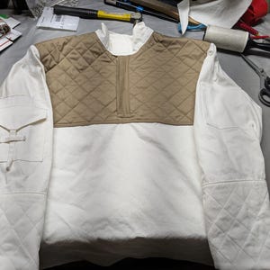 Può includere: Giacca trapuntata bianca e beige con collo alto e zip a un quarto. La giacca presenta un pannello beige trapuntato sul petto e sulla schiena superiore, con maniche e corpo bianchi. Ha una tasca sulla manica sinistra.