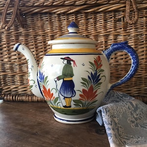 May include: A hand-painted ceramic teapot with a lid, featuring a design of a person in traditional attire. The teapot has a white base with blue, orange, and green floral accents, a yellow band, and a blue handle. The teapot is on a wooden surface.