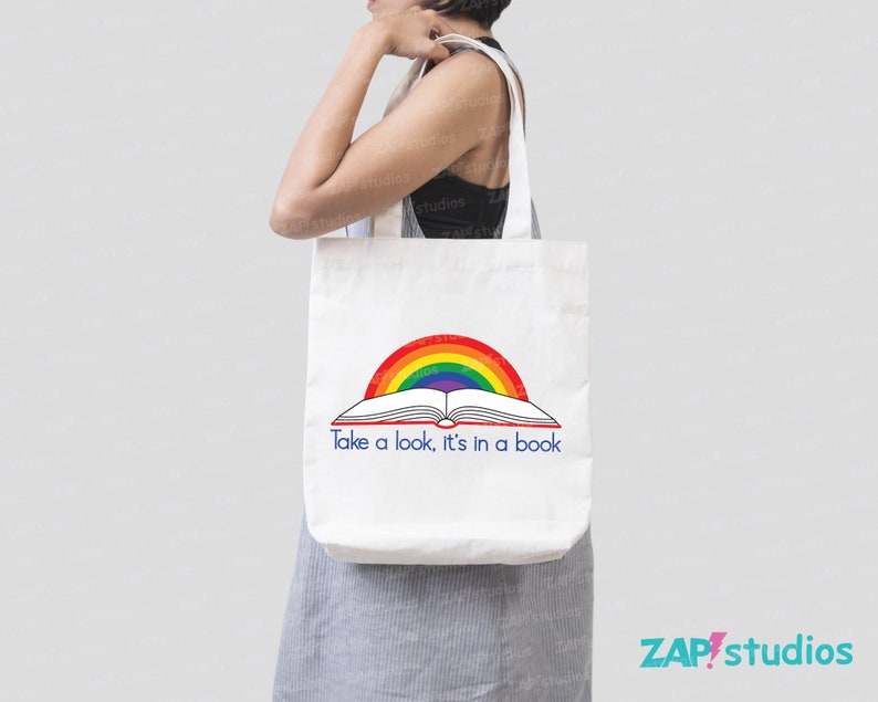 May include: A white tote bag with a rainbow and an open book design. The text on the bag reads "Take a look, it's in a book."