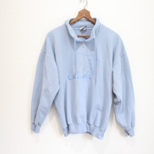 May include: A light blue long-sleeve sweatshirt featuring a quarter-zip collar with a drawstring and a graphic of a bear with the text Alaska printed in a darker blue shade on the front, hanging on a wooden hanger against a plain white background.