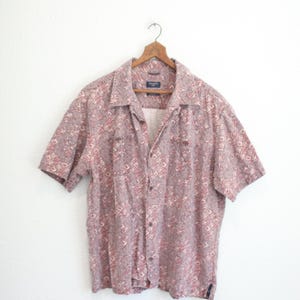 May include: Short-sleeved button-down shirt with a geometric pattern in shades of red, white, and gray. The shirt is hanging on a wooden hanger against a white background. Features a collared neckline and two chest pockets.