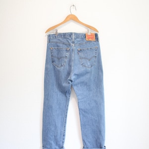 May include: Light blue denim jeans hanging on a wooden hanger. The jeans feature a classic five-pocket design, a red brand tab on the right back pocket, and a tan leather-like patch on the back waistband with visible red and black brand text.