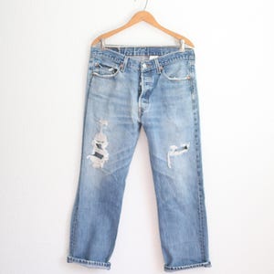 May include: Light blue denim jeans with distressed details, hanging on a wooden hanger. The jeans have a button fly, front pockets, and a relaxed fit. The cuffs are rolled up, and there are rips on the knees.