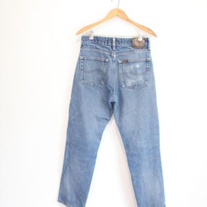 May include: A pair of light blue denim jeans shown from the back, featuring two rear pockets, a brown leather brand patch on the waistband, and a classic straight-leg cut. The jeans are suspended on a wooden hanger against a plain white background.