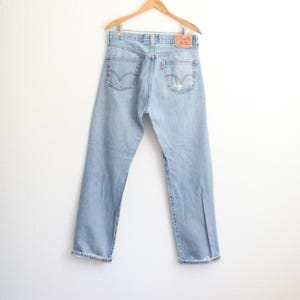 May include: Light blue denim jeans hanging on a wooden hanger. The jeans feature two back pockets with a signature arcuate stitch design, a tan leather-like patch on the waistband, and a small red tab on the right pocket. The hems are cuffed.