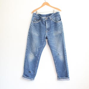 May include: Blue denim jeans featuring a classic five-pocket design, a button fly, and a medium wash with subtle fading on the thighs. The jeans have a straight-leg cut with rolled cuffs at the bottom hem and are displayed on a wooden hanger.