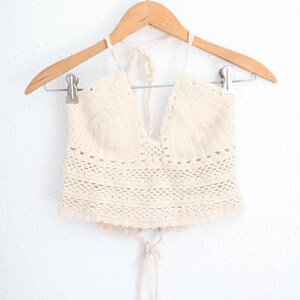 May include: A cream-colored crochet halter top featuring a deep V-neckline, intricate openwork stitch patterns, and adjustable tie closures at the neck and back. The garment is displayed on a wooden hanger with metal clips for support.