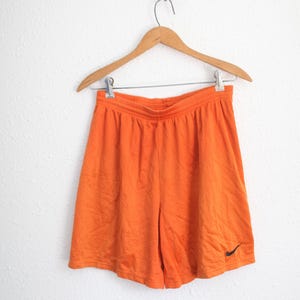 May include: Bright orange athletic shorts featuring a mesh fabric construction with an elasticized waistband and a small black Nike swoosh logo embroidered on the lower left leg, suspended from a wooden hanger with metal clips.