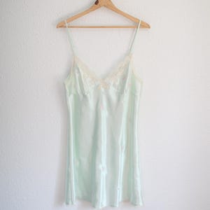 May include: A light mint green satin slip dress featuring thin spaghetti straps and a V-neckline trimmed with delicate cream-colored lace. The fabric has a subtle sheen and is decorated with small, scattered floral embroidery throughout the garment.