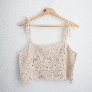 May include: A cream-colored crochet crop top featuring a repeating hexagonal pattern with intricate star-like motifs. The garment has thin, adjustable tie-up shoulder straps and a scalloped bottom hem, all displayed on a wooden hanger.