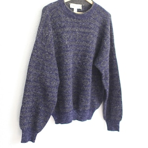 May include: A vintage crew neck sweater in a speckled navy blue and olive green pattern. The sweater has long sleeves and a ribbed hem and cuffs. It is hanging on a wooden hanger against a white background.